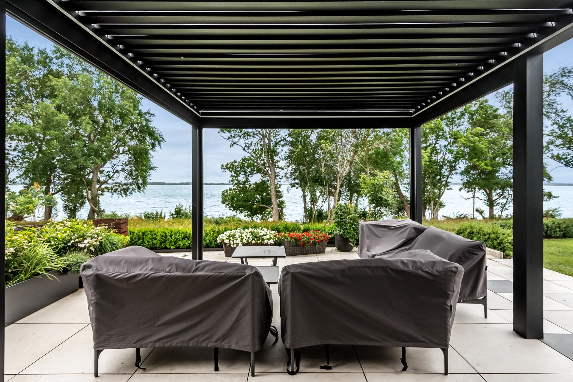 Sittgrupp under en svart pergola med lamelltak och utsikt över sjön